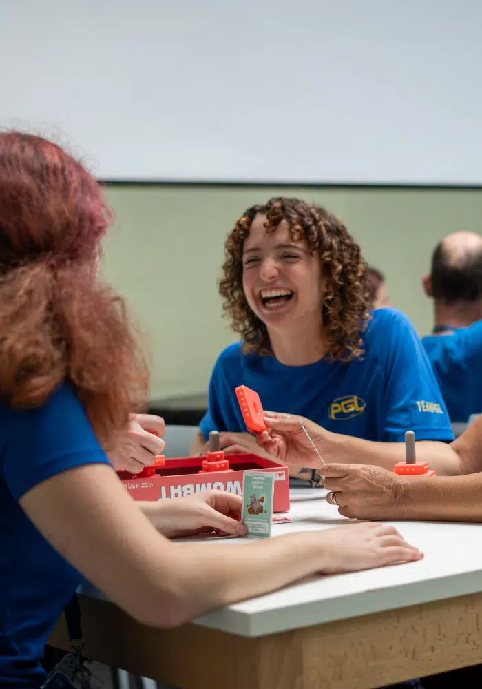 People in blue shirts are sitting around a table, playing a word game with red buzzers and cards. One person is laughing.