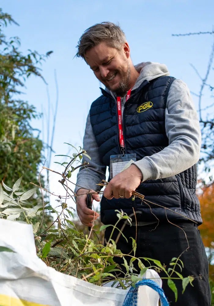 A man wearing a vest and hoodie trims branches with pruning shears outdoors. He stands next to a large bag filled with clippings.