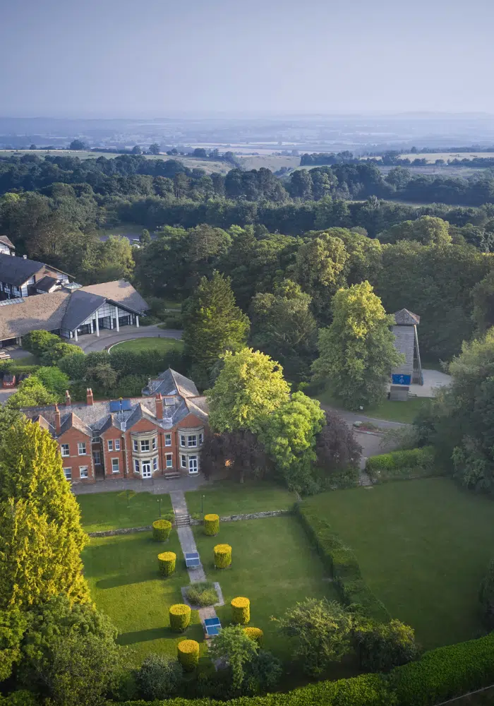 Aerial view of a large brick house with a manicured garden, surrounded by trees and countryside under a clear sky.