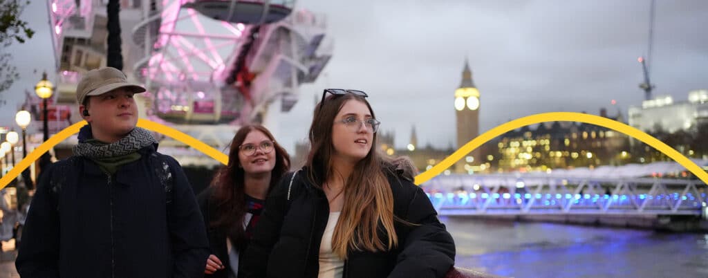 Three people stand near the London Eye and Big Ben, with a cityscape in the background during twilight.
