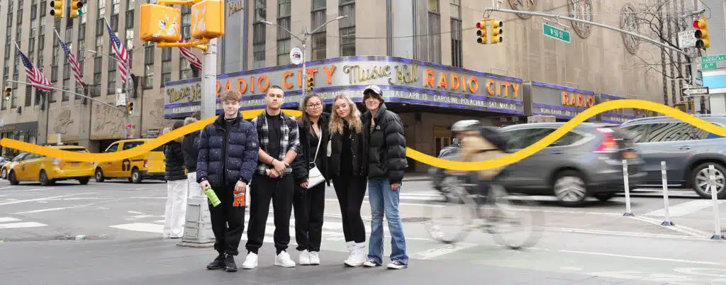 Five people stand on a city street in front of Radio City Music Hall. Traffic and a cyclist pass by.