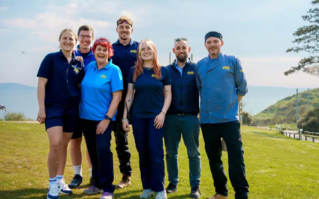 A group of seven people in PGL uniforms posing outdoors on a grassy area with a view of the ocean in the background.