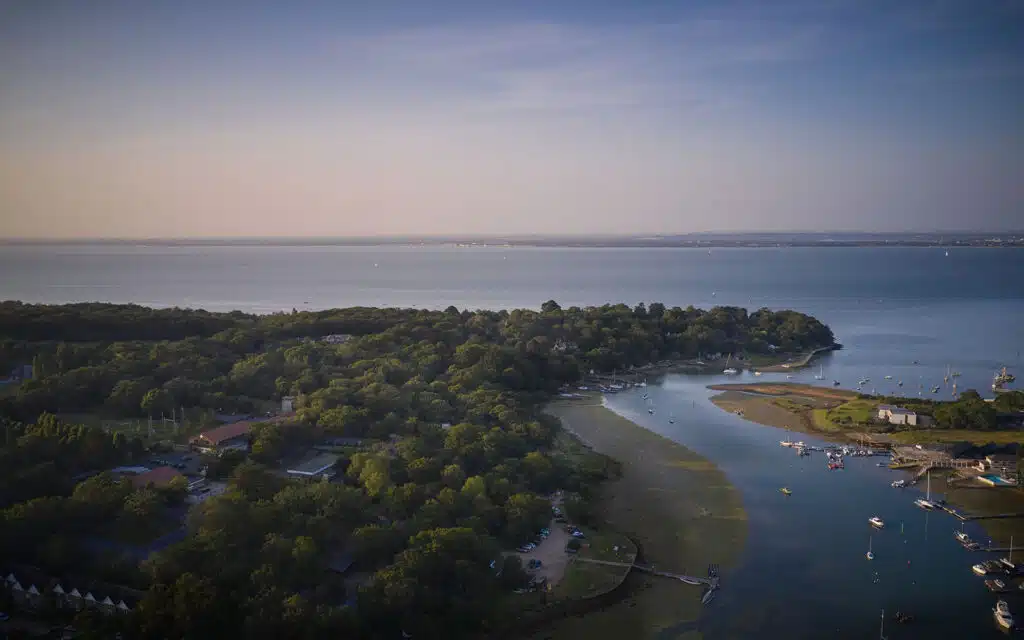 Aerial view of PGL Little Canada with dense greenery, a winding river leading to the sea, and boats docked along the shore under a clear sky.