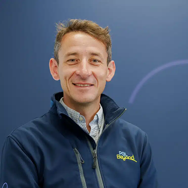 A person with short brown hair wearing a dark blue jacket and a light shirt, standing against a plain dark background.