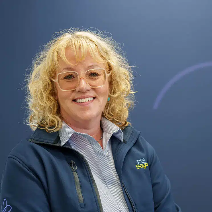 Smiling person with curly hair and glasses wearing a blue jacket, standing against a blue background.