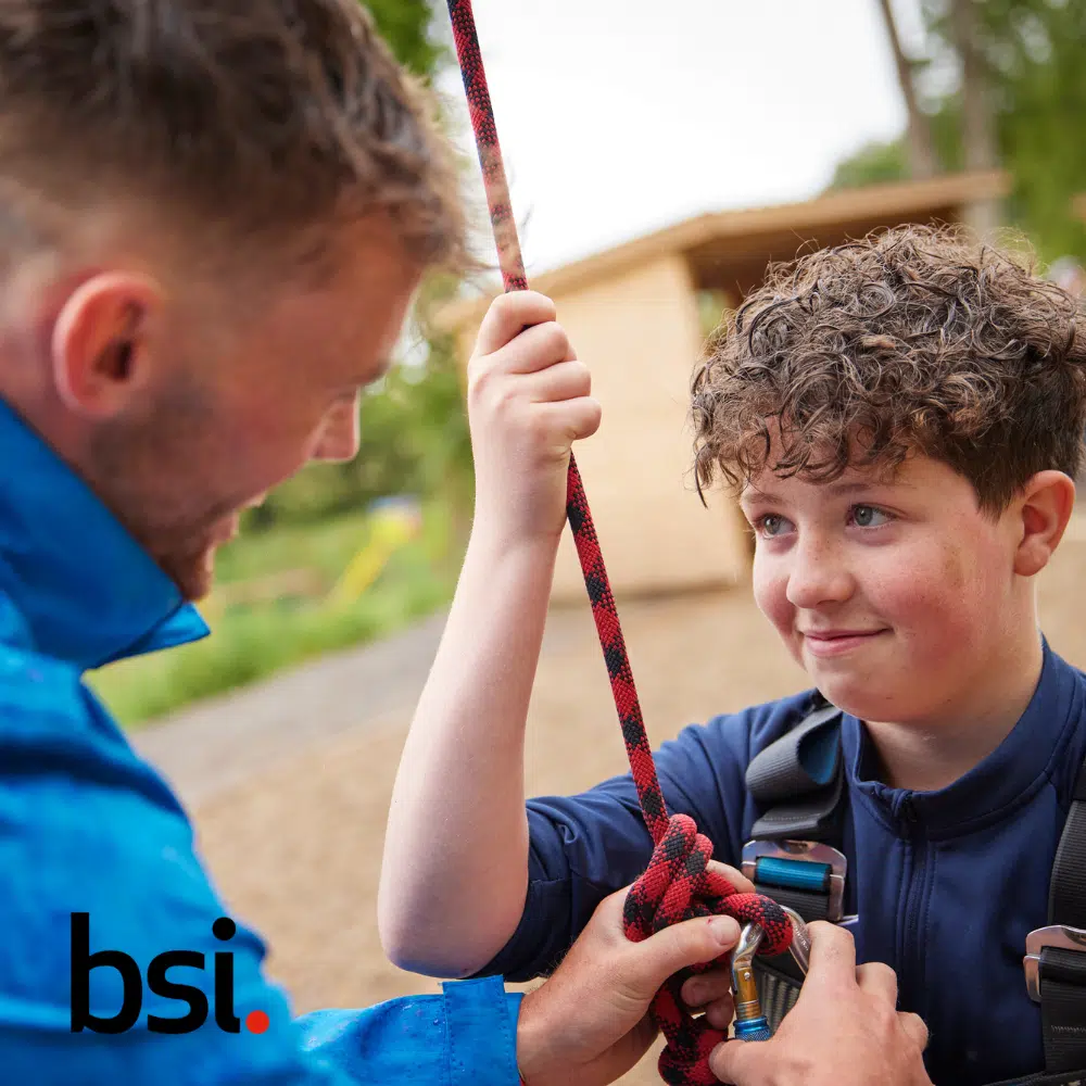 A man helps a child secure a rope harness outdoors. The child looks up at the man. The BSI logo is in the bottom left corner.