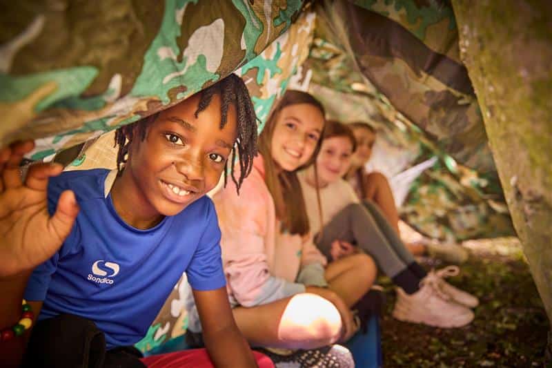 Children sitting under a camouflage tarp, smiling at the camera.