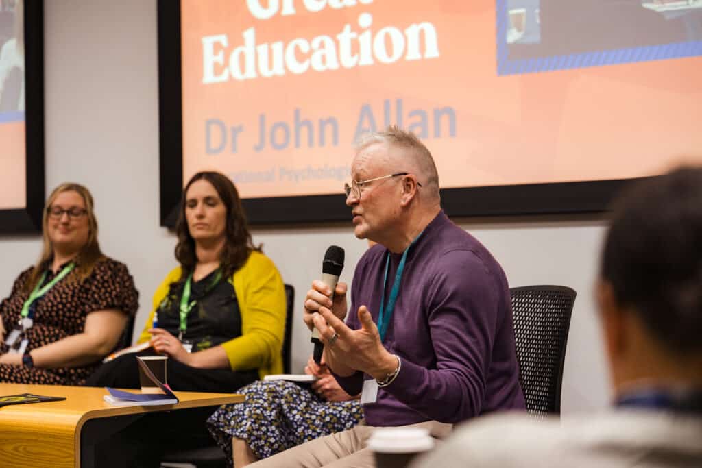 A person speaks into a microphone during a panel discussion, with two others seated beside them. A presentation screen is visible in the background.