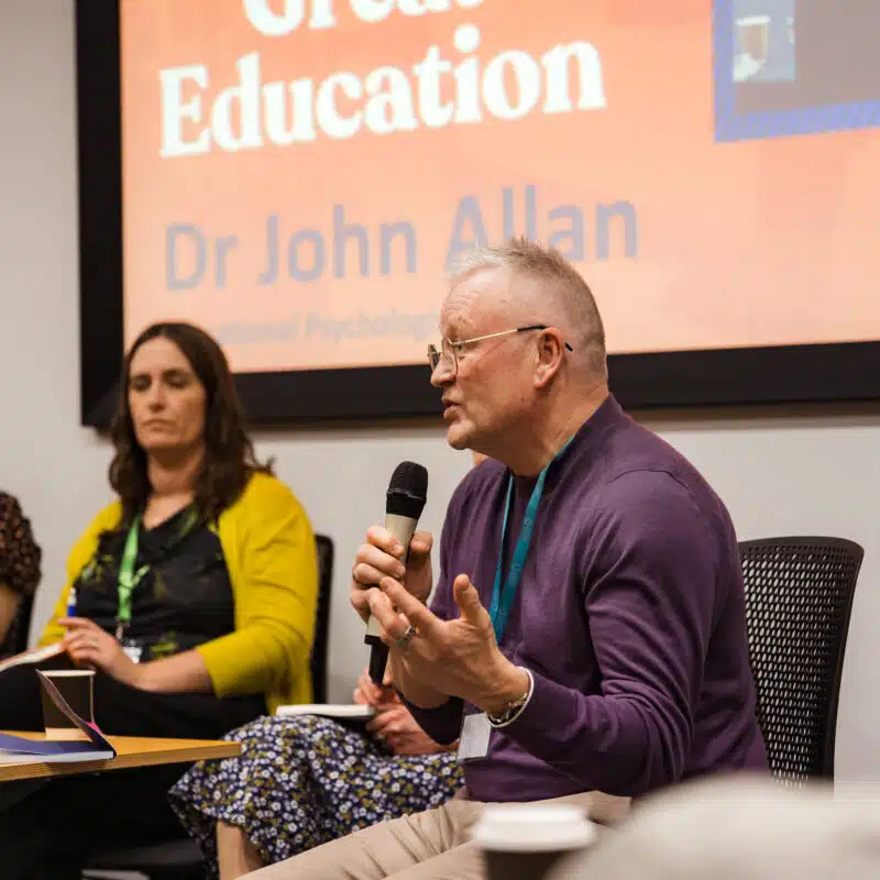 A person speaks into a microphone during a panel discussion, with two others seated beside them. A presentation screen is visible in the background.