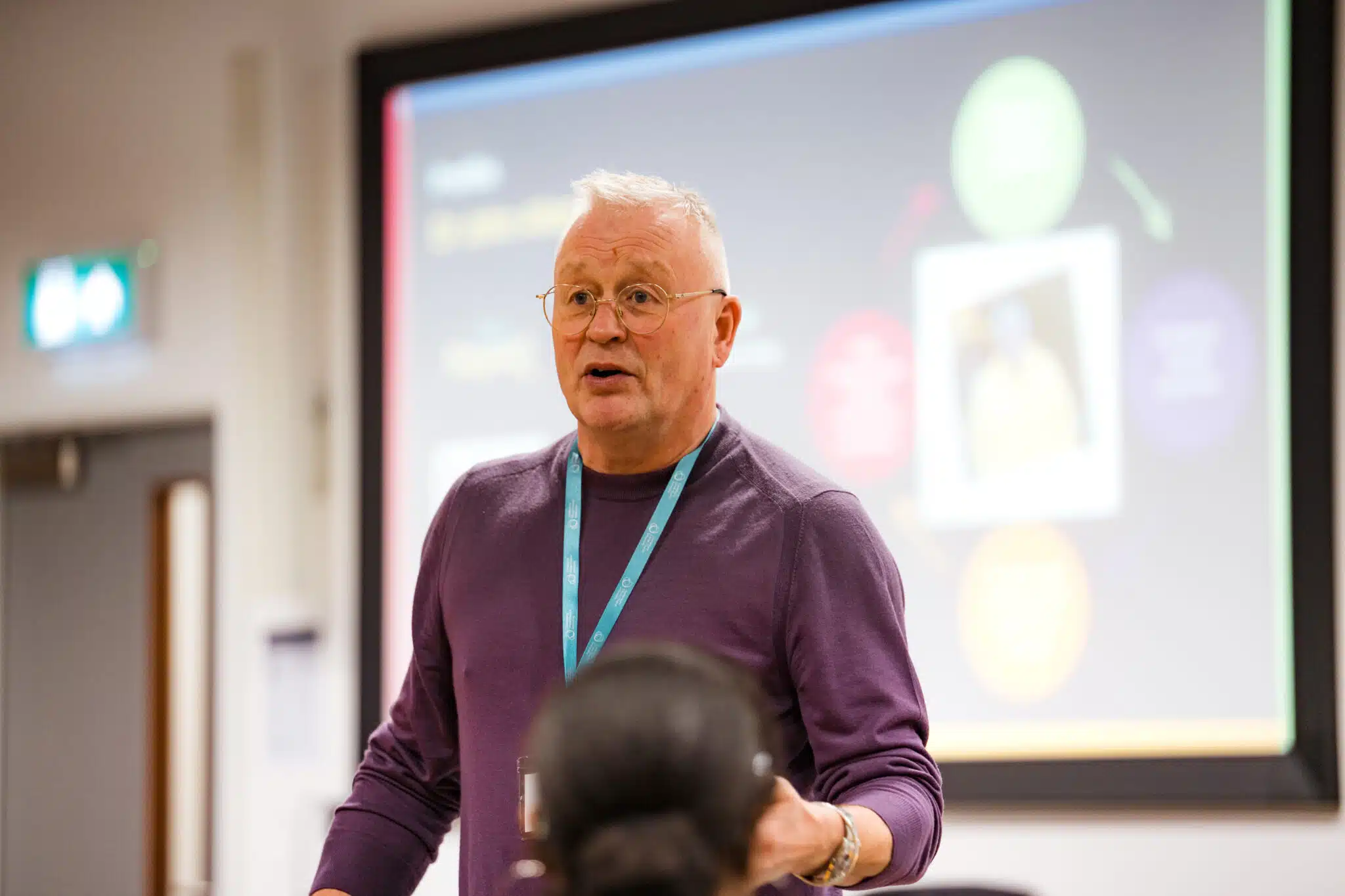 Man with glasses and purple shirt speaking in front of a blurred presentation screen in a classroom setting.
