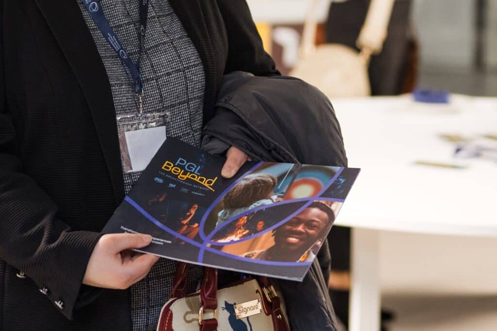 Person holding a booklet titled "PGI Beyond," featuring an image of a smiling person on the cover, standing indoors near a white table.