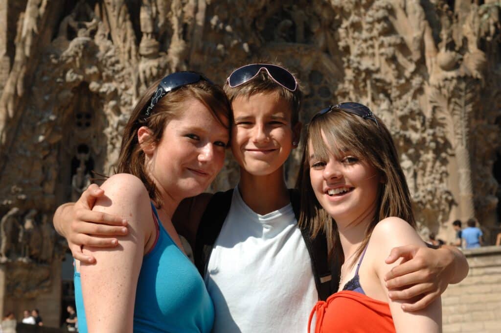 Three smiling individuals pose together in front of a detailed stone facade, with one person in the center wrapping their arms around the others.