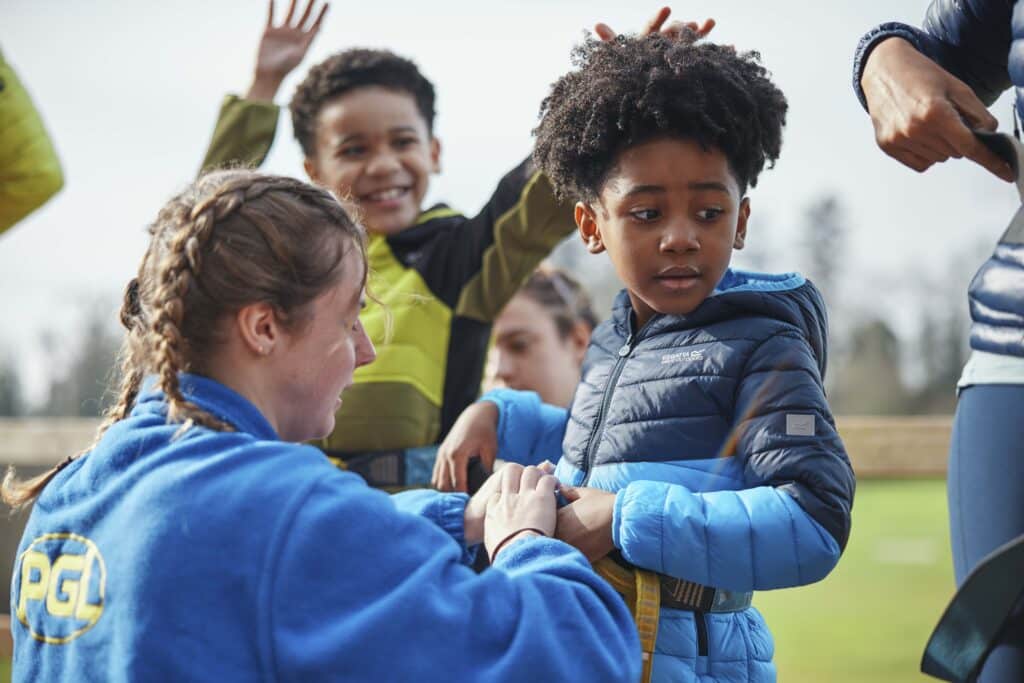 A woman helps a child adjust a harness outdoors, while another child stands nearby raising their arms.