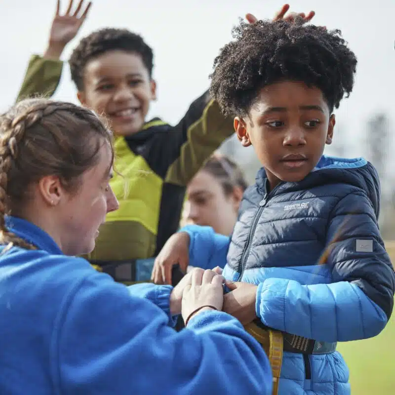 A woman helps a child adjust a harness outdoors, while another child stands nearby raising their arms.