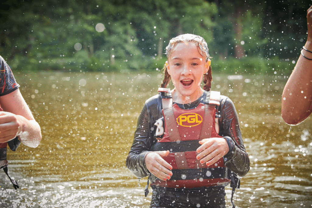 A girl wearing a red PGL life jacket stands in shallow water outdoors, splashed by water, with greenery blurred in the background.