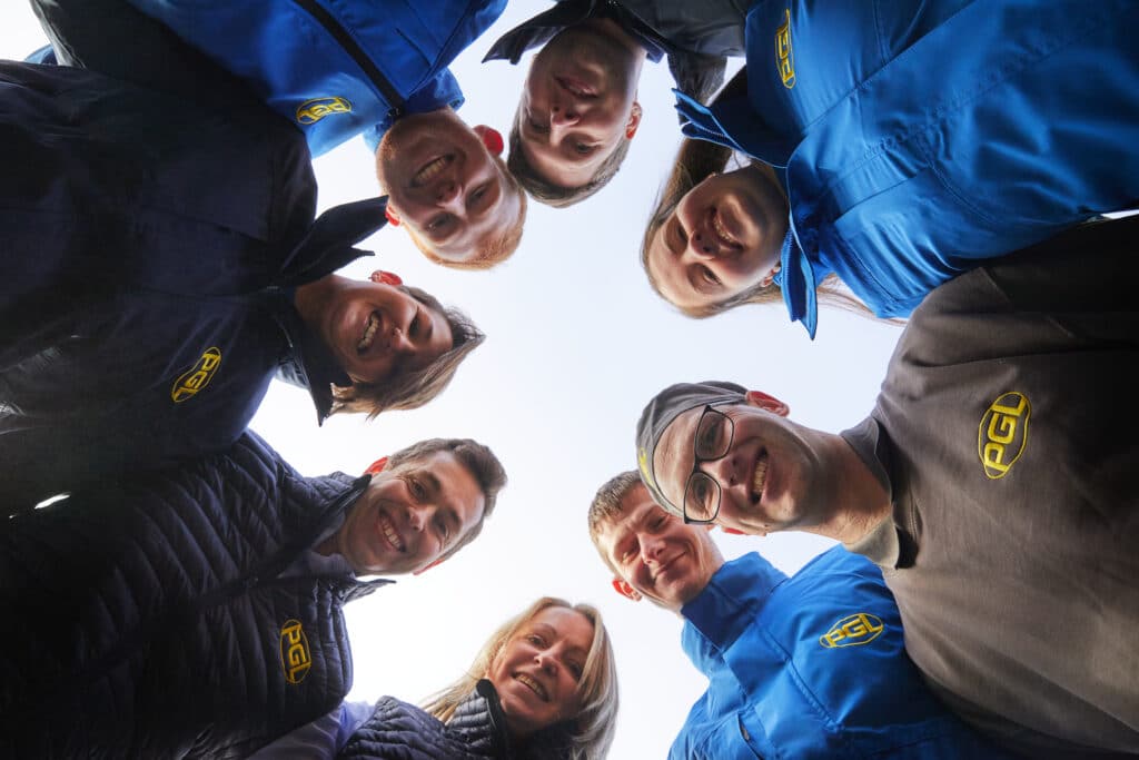 Eight people wearing PGL-branded jackets and shirts stand in a circle, looking down at the camera and smiling, with a clear sky background.