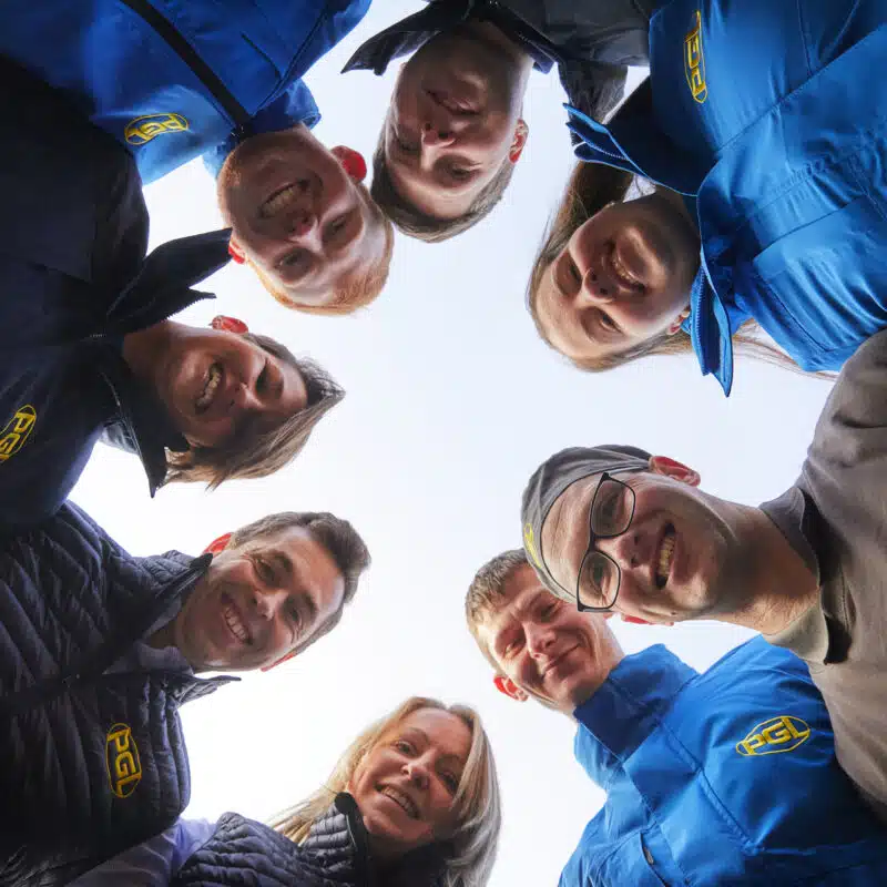 Eight people wearing PGL-branded jackets and shirts stand in a circle, looking down at the camera and smiling, with a clear sky background.