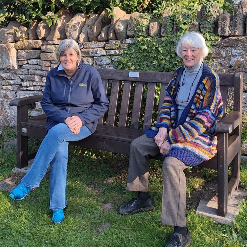 Two people sit on a wooden bench in a garden, next to a stone wall and metal gate. One wears a blue jacket and jeans, the other a colorful sweater and gray pants. Grass and trees surround them.