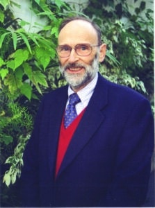 A man with a beard and glasses, wearing a navy blazer, red sweater, and blue tie, standing in front of green foliage.