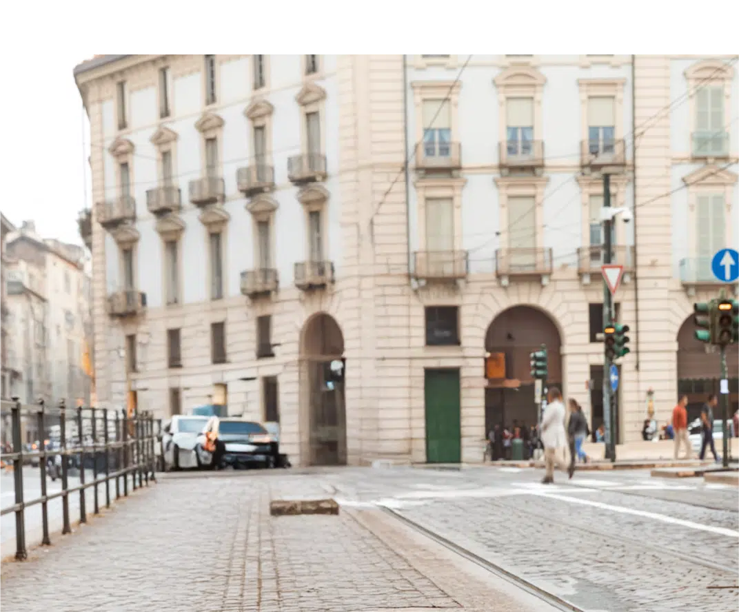 A city street with tram tracks, traffic lights, and pedestrians crossing near a historic, multi-story building on a cloudy day.