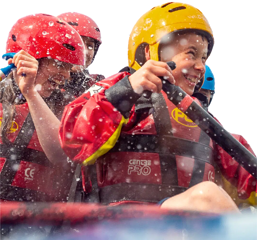 Children wearing helmets and life jackets paddle a boat, water splashing around them as they smile and enjoy the activity.