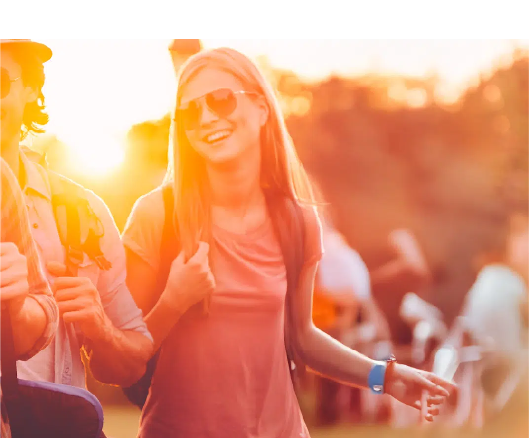 A woman wearing sunglasses and a backpack smiles while walking outdoors in the sunlight with other people in the background.