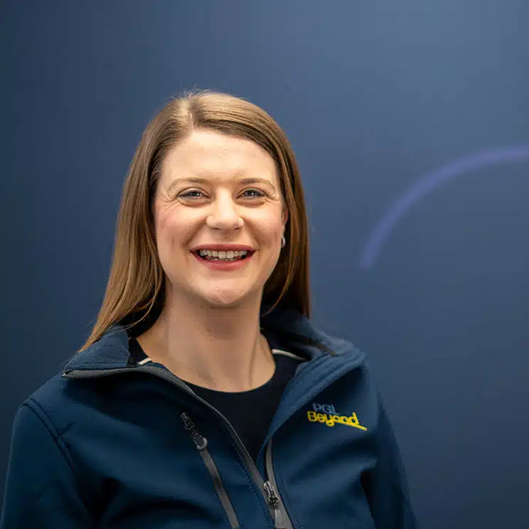 A woman with straight, light brown hair smiles at the camera, wearing a navy zip-up jacket with a "PGE Beyond" logo, standing in front of a plain blue background.