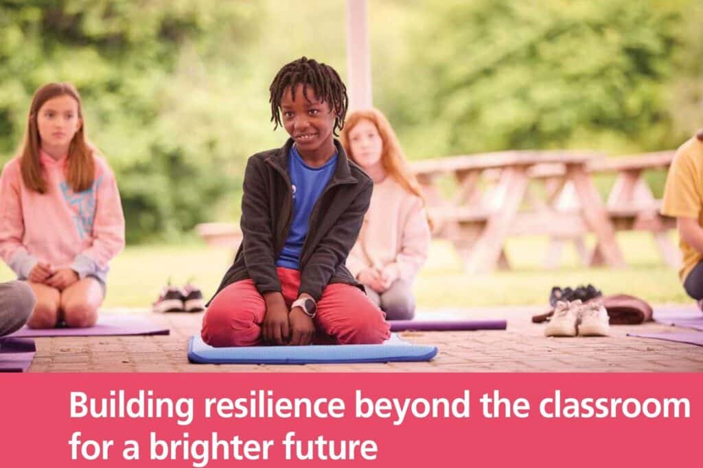 Several children sit on mats outdoors, with one child in the center looking at the camera. Text reads: "Building resilience beyond the classroom for a brighter future.