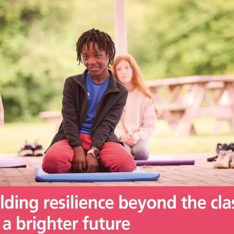 Several children sit on mats outdoors, with one child in the center looking at the camera. Text reads: "Building resilience beyond the classroom for a brighter future.