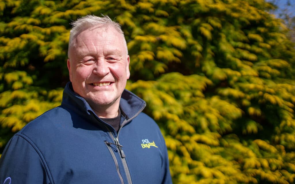 A man with short blonde hair wearing a blue jacket stands outdoors in front of a green and yellow leafy bush, smiling at the camera.