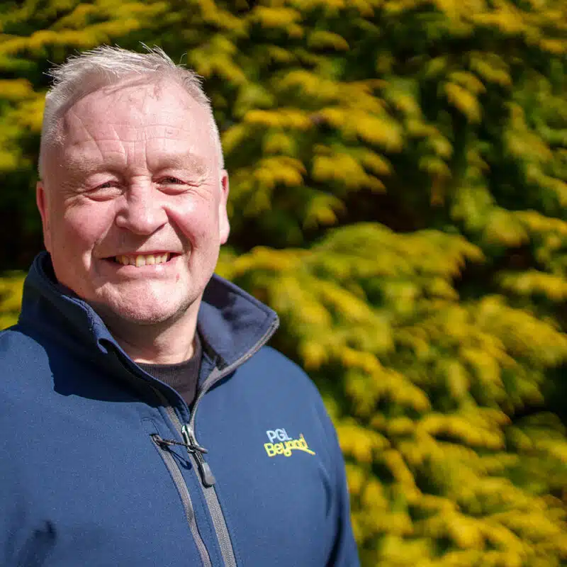 A man with short blonde hair wearing a blue jacket stands outdoors in front of a green and yellow leafy bush, smiling at the camera.