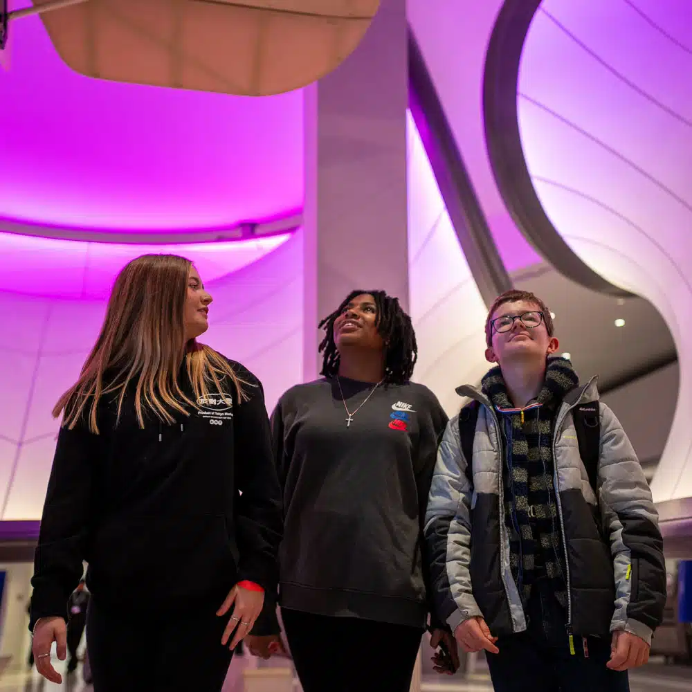Three people stand indoors under curved, purple-lit architecture, looking upward and appearing engaged with their surroundings.