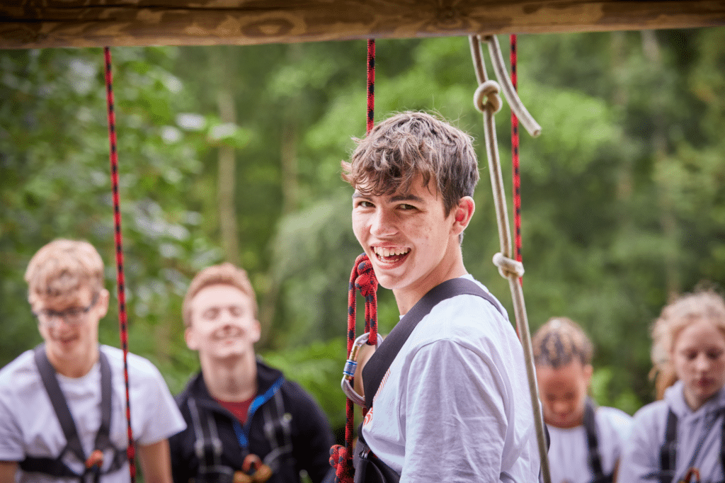 A young person wearing a harness smiles at the camera, with other people in harnesses and trees in the background.