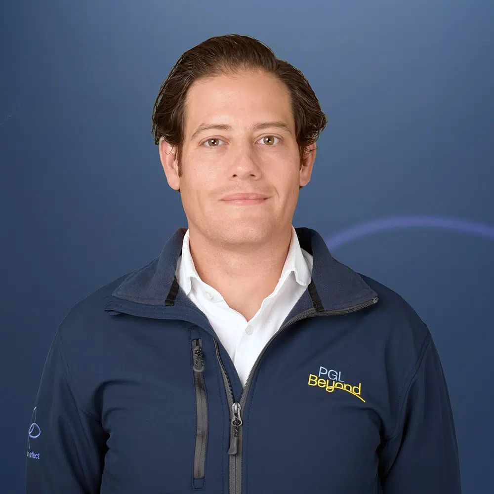 A man with short brown hair wears a white shirt and a navy jacket with a "PGL Beyond" logo, standing in front of a blue background.