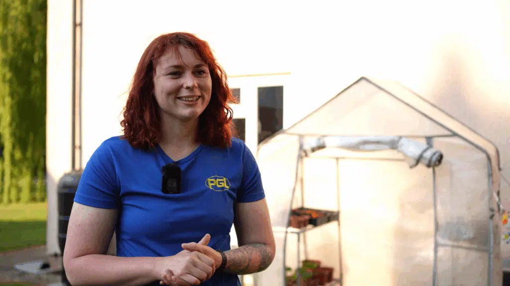 A person with red hair, wearing a blue PGL shirt and a microphone, stands outside near a small greenhouse and a building wall.