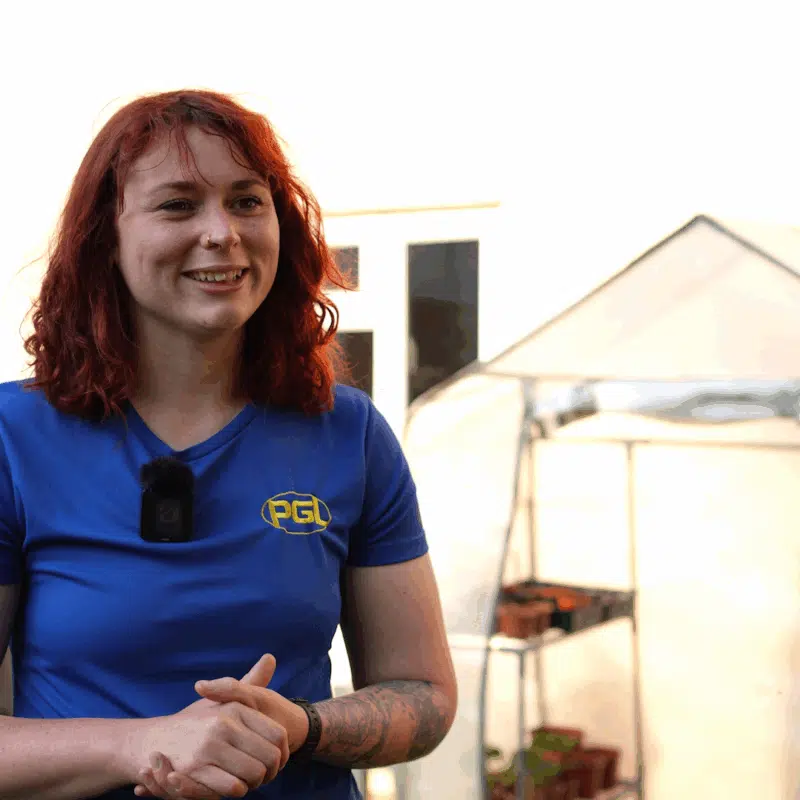A person with red hair, wearing a blue PGL shirt and a microphone, stands outside near a small greenhouse and a building wall.