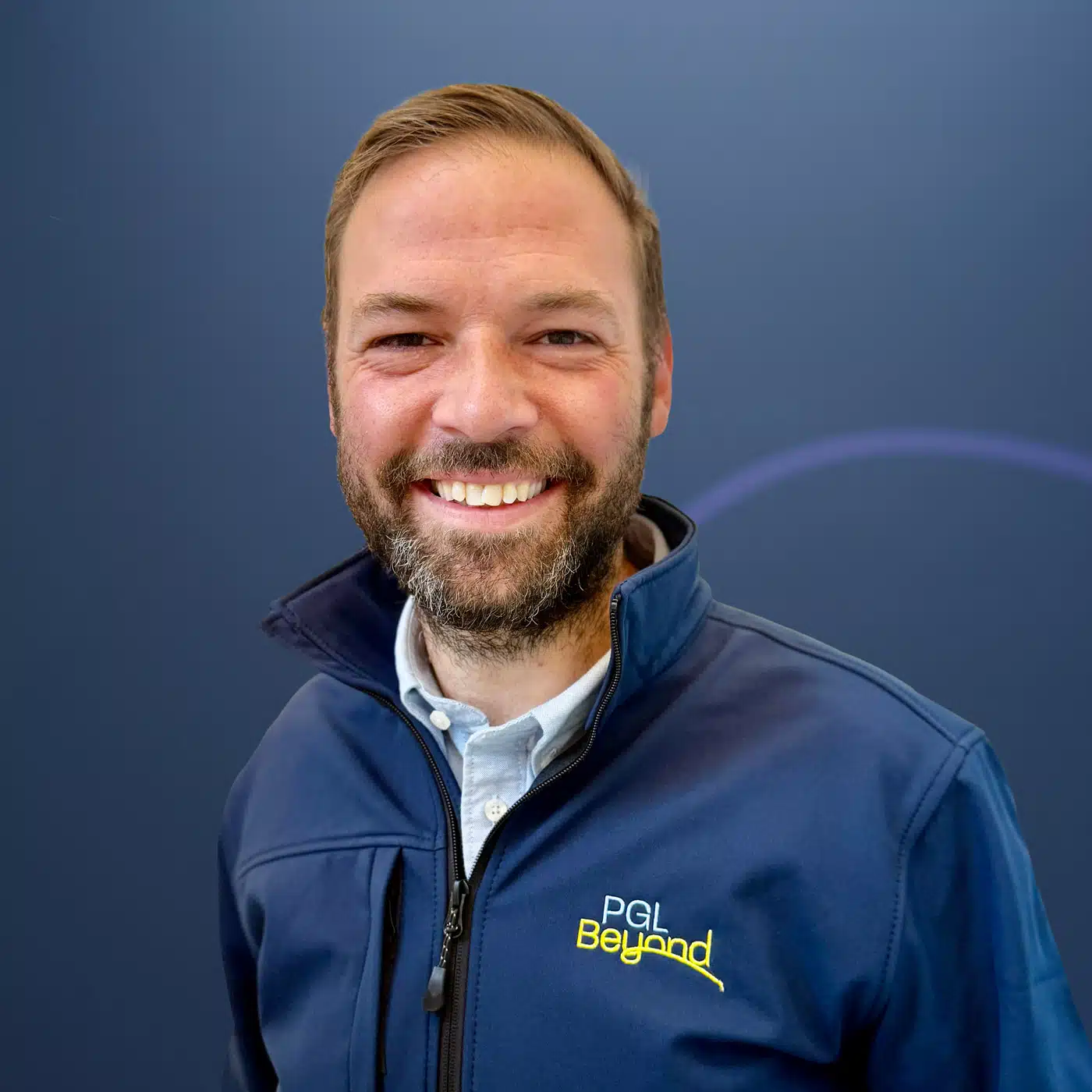 A man with a beard smiles at the camera, wearing a blue jacket with a "PGL Beyond" logo, standing in front of a plain blue background.
