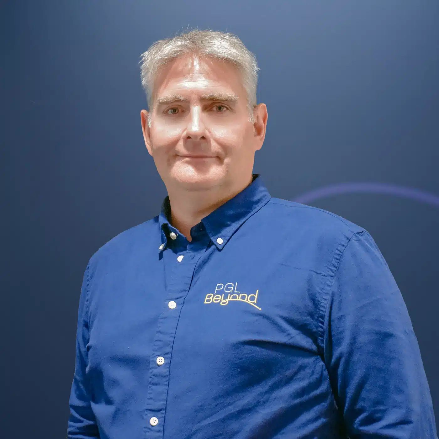 A man with short gray hair wearing a blue button-up shirt with a "PGL Beyond" logo stands in front of a plain blue background.