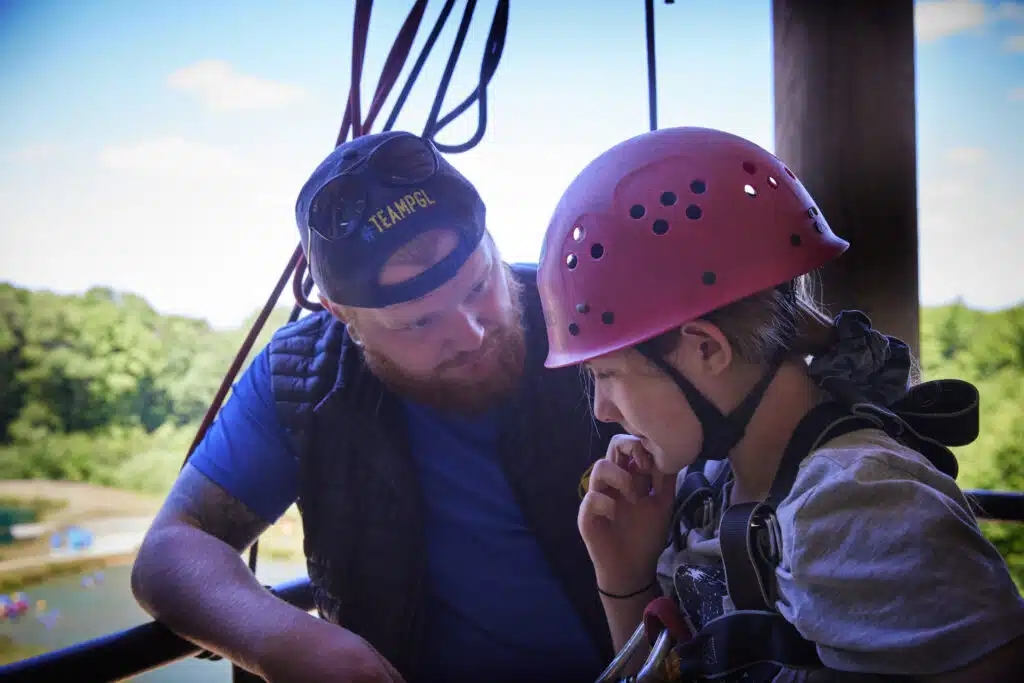 An adult wearing a cap and vest talks to a child in a helmet and harness, standing together on an outdoor platform with trees in the background.
