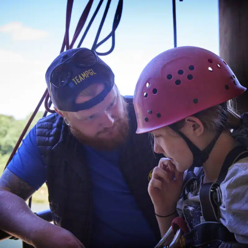 An adult wearing a cap and vest talks to a child in a helmet and harness, standing together on an outdoor platform with trees in the background.