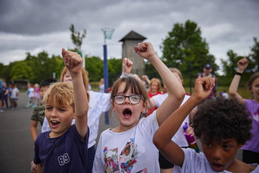 A group of children outdoors raise their fists and shout, appearing energetic and enthusiastic, with trees and playground equipment visible in the background.