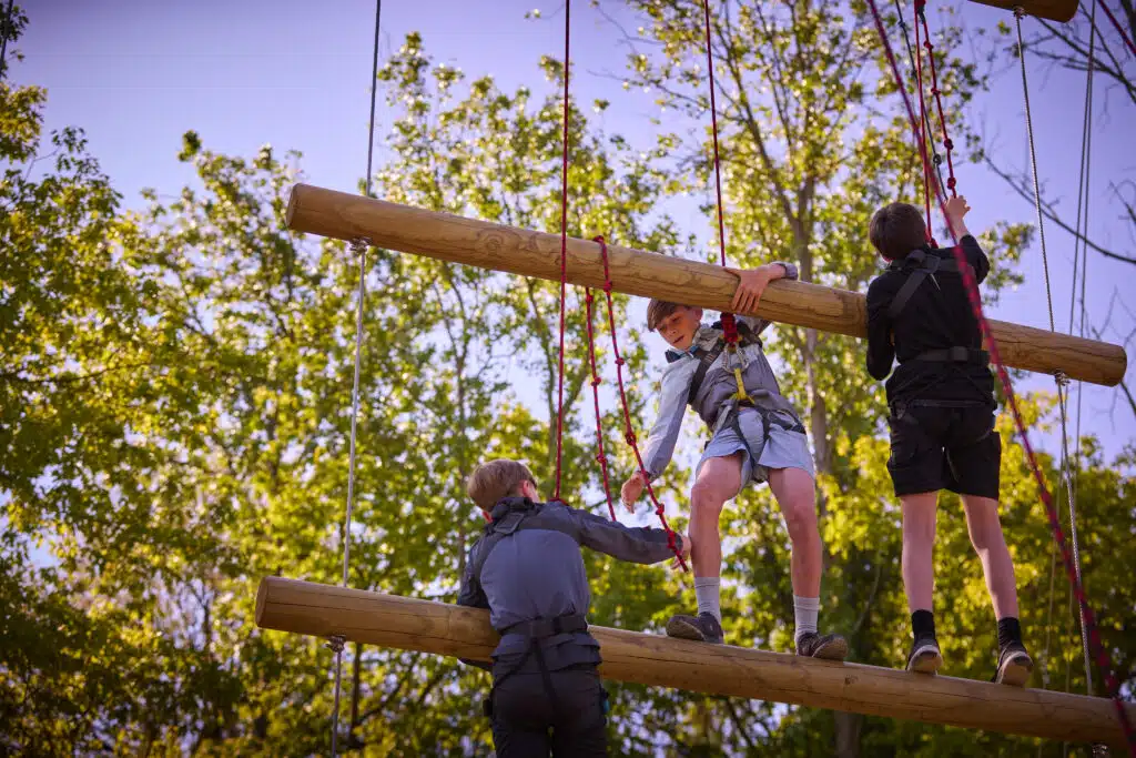 Three children wearing harnesses climb and balance on elevated wooden beams in an outdoor ropes course, surrounded by trees.