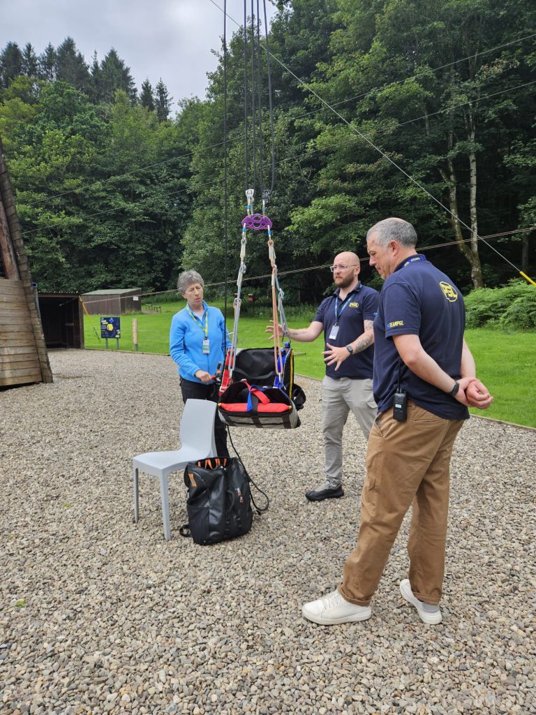 Three people stand outdoors beside a harness and ropes suspended between trees, with a white chair and backpack nearby on a gravel surface.