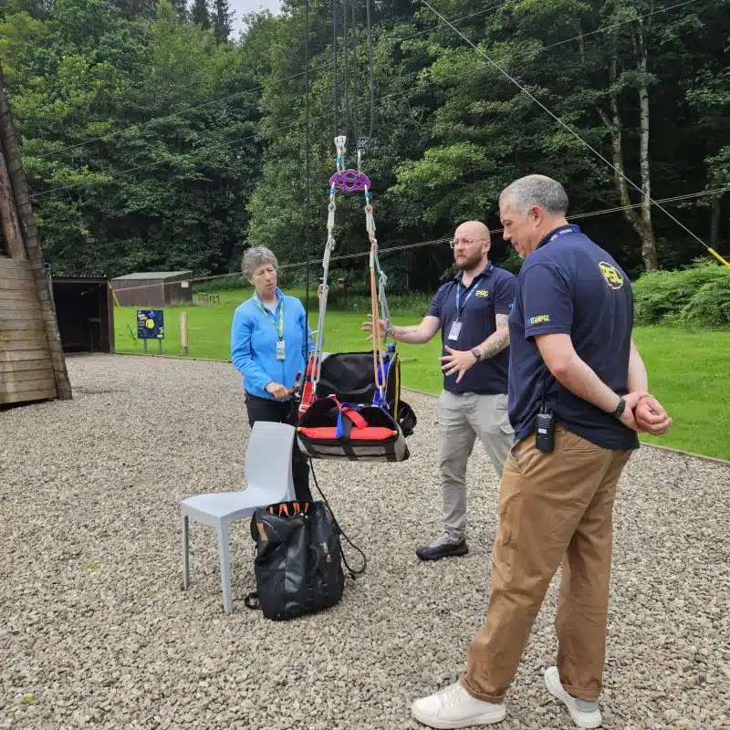 Three people stand outdoors beside a harness and ropes suspended between trees, with a white chair and backpack nearby on a gravel surface.