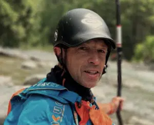 A person wearing a helmet and blue life jacket holds a paddle near a river with trees in the background.