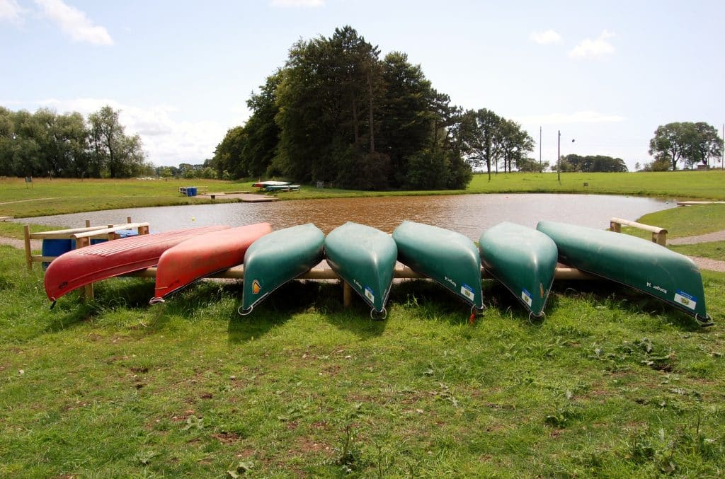 Seven canoes, mostly green with one red, are stored upside down on wooden racks by a small pond in a grassy park with trees in the background.