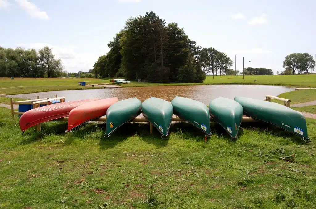 Seven canoes, mostly green with one red, are stored upside down on wooden racks by a small pond in a grassy park with trees in the background.