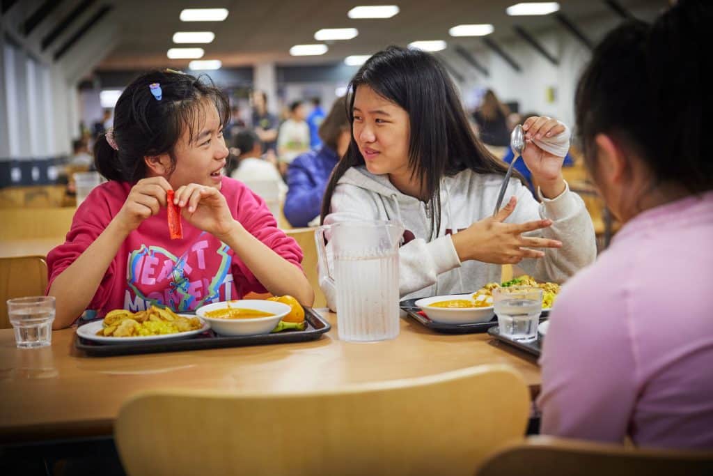Three girls sit at a cafeteria table with trays of food, talking and smiling. A water pitcher and glasses are on the table. The background shows a busy dining hall.