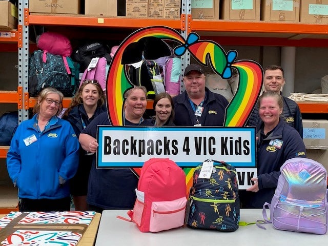 A group of seven people stand in a warehouse holding a "Backpacks 4 VIC Kids" sign, with colorful backpacks displayed on the table in front of them.