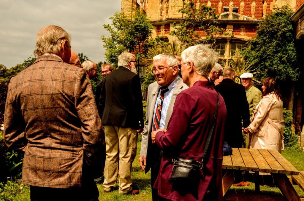 A group of older adults dressed in formal attire converse outdoors in a garden near a brick building, with a wooden picnic table visible in the foreground.